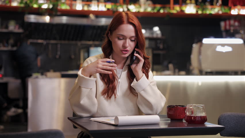 Caucasian woman with long red hair sitting in cafe. Talking on smartphone and pointing at notebook. Focused female freelancer discussing about work while reviewing notes at table with drink.