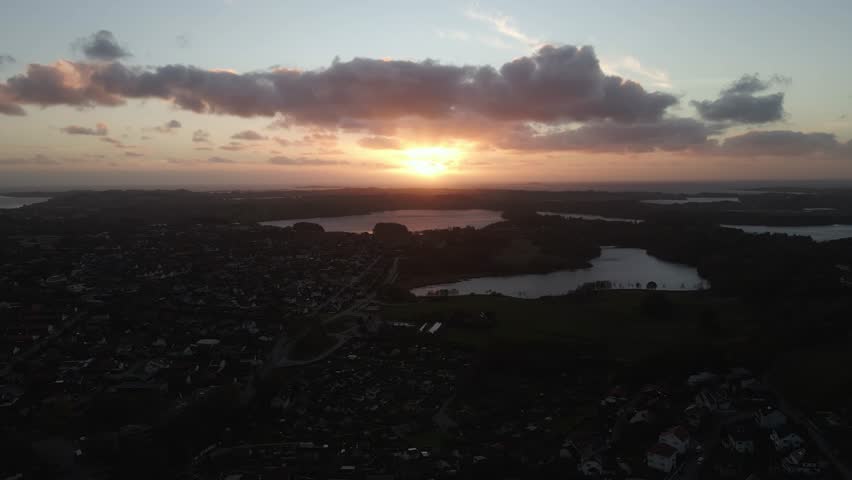 Aerial drone footage showing lakeside suburbs under a cloudy sunset. The scene captures soft evening light reflecting on the water, with suburban homes and trees lining the shore, creating a calm and 