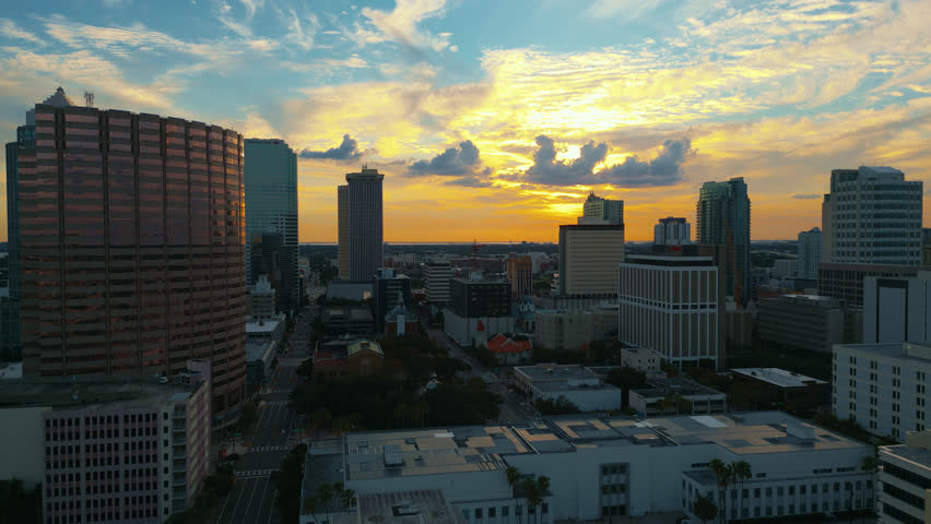Tampa skyline at sunset with silhouetted buildings and golden light reflecting off rooftops, aerial pullback.