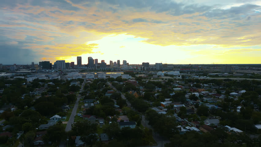 Aerial sunrise or sunset golden hour establishing shot over a neighborhood near downtown Tampa with glowing clouds and skyscrapers in the distance.