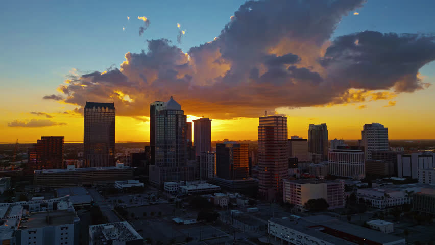 Aerial drone tracking shot of the Tampa, FL skyline at golden hour. As the camera tracks right the sun peaks out from behind the tall buildings and subtle flares are created.