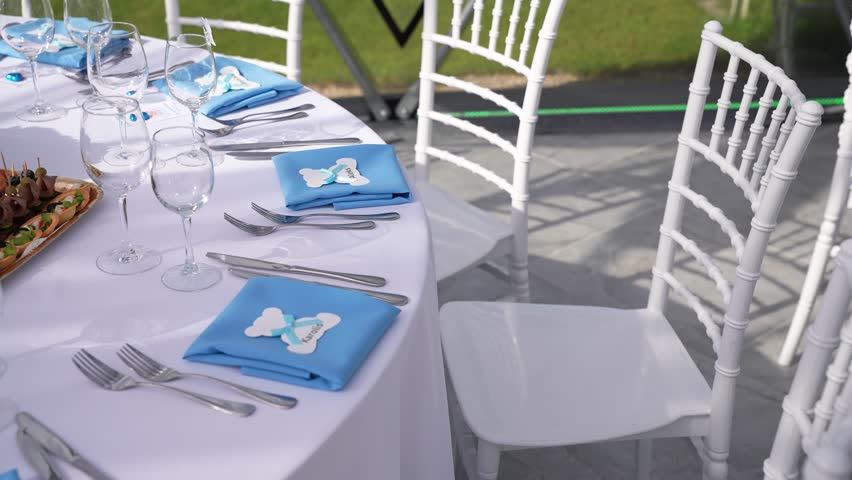 Elegant christening table setup with blue napkins, personalized tags, white chairs, and glassware arranged for a formal baby boy celebration in a bright, airy venue.