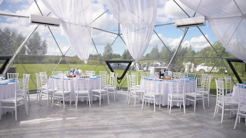 Elegant wedding reception setup inside a transparent dome with white chairs and round tables, overlooking a scenic green landscape through large glass panels.