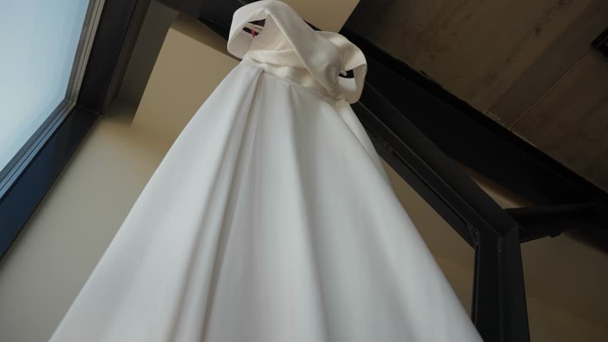 Close-up of a white satin wedding dress hanging indoors, showcasing its elegant neckline and soft flowing fabric, perfect for a romantic bridal moment, zooming in.