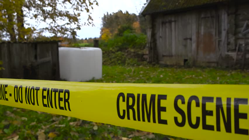 Slow tracking shot moves along yellow crime scene tape stretched across the front of an old blurred countryside wooden house. Criminal investigation site around rural barn shed during overcast autumn.