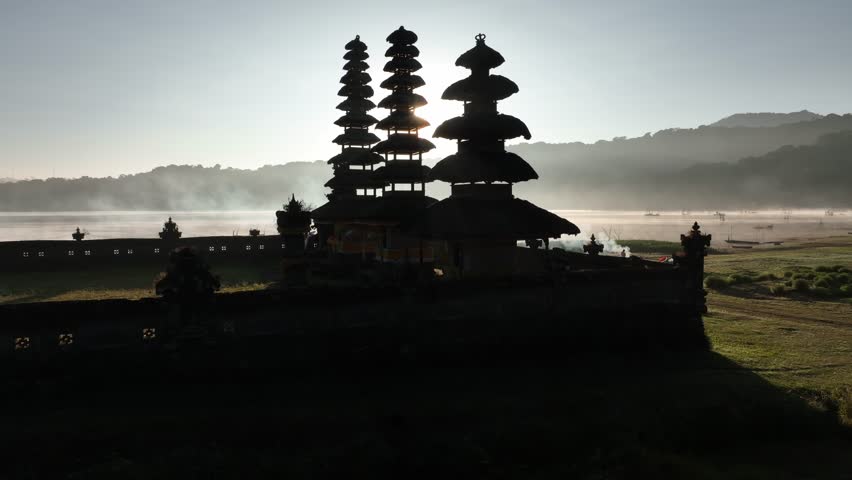 Aerial view of Pura Ulun Danu Tamblingan, a temple silhouette against the sun and misty lake, creating a serene landscape, Pura Ulun Danu Tamblingan, Bali, Indonesia.