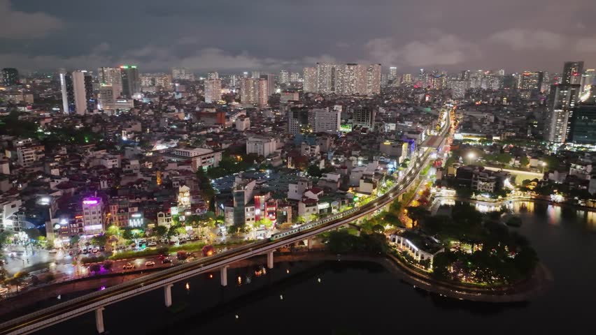 Night aerial view of Hanoi with metro line and city lights reflecting on Hoang Cau lake.