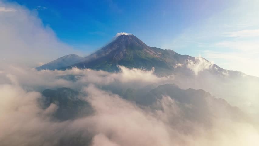Aerial view of Gunung Merapi Mountain peak piercing through a sea of swirling clouds, its slopes a stark contrast to the lush green forests below, Yogyakarta, Indonesia.