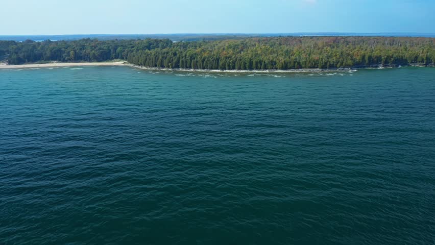 The deep blue surface of Lake Michigan ripples gently toward Door County’s forested shoreline, where dense green trees edge the coast beneath clear afternoon light.