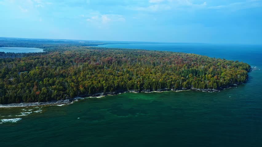 The forested tip of Door County reaches into Lake Michigan, its trees glowing with early autumn hues above turquoise waves and rocky shores under a clear blue sky.