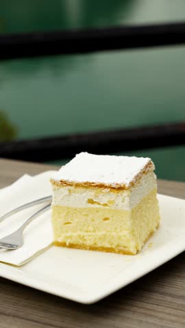 Bled cream cake Kremšnita sits on a cafe plate with Lake Bled beyond, gentle right vertical orbit.
