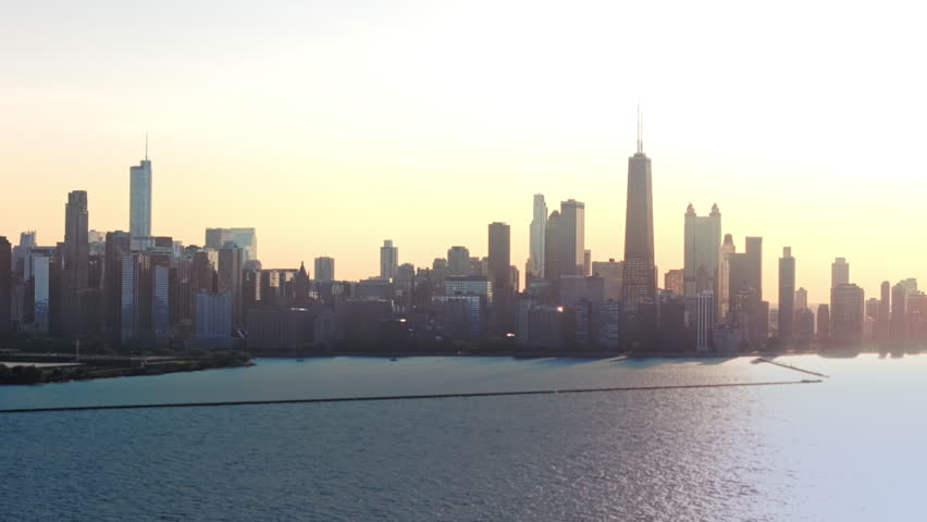 A 70mm, cinematic drone shot slowly rotating from left to right, far over Lake Michigan, as the setting sun casts a warm glow over a skyline of skyscrapers and the sprawling metropolis of Chicago, IL.