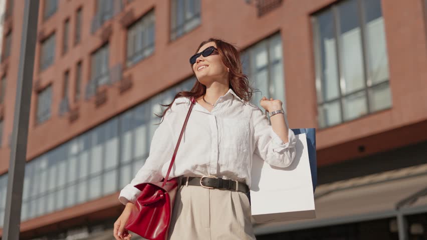Smiling Caucasian woman in white shirt holding shopping bag. Wearing sunglasses and looking up with joy. Carrying red handbag while enjoying sunny day outdoors. Modern buildings in background.