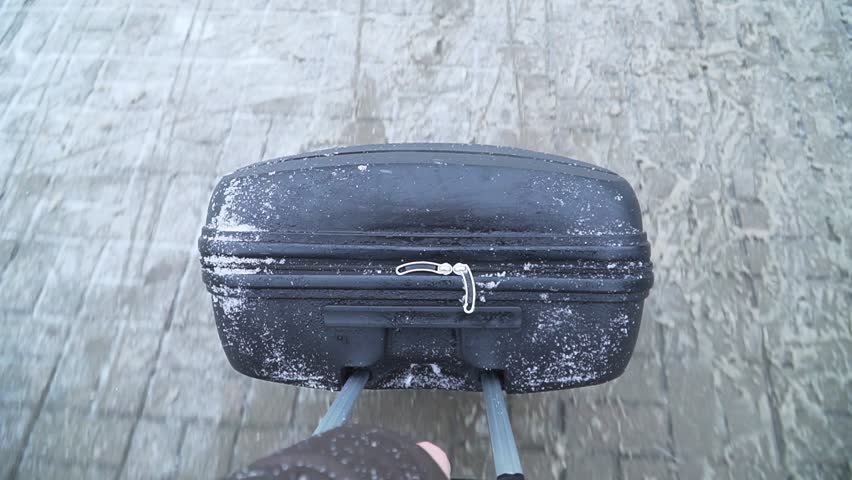 Close-up view of a black suitcase being pulled across a snowy surface, showcasing the texture of snow and the suitcase's details, emphasizing movement and travel in winter conditions