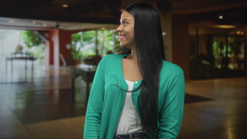 Woman wearing green cardigan over white top smiles softly with long black hair draped over shoulder in building; serenity.