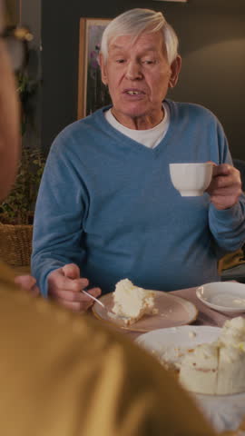 Vertical over the shoulder view of smiling senior man speaking with friend while having tea at home birthday party