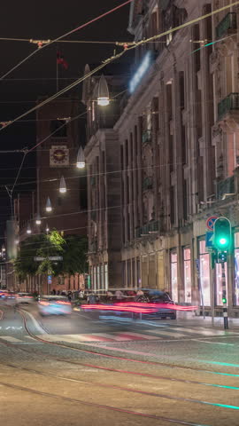 Electric tram on Damrak Street heading from Dam Square to Centraal Station in a timelapse at night. Traffic moves on the road while people wait at the tram stop. Amsterdam, Netherlands