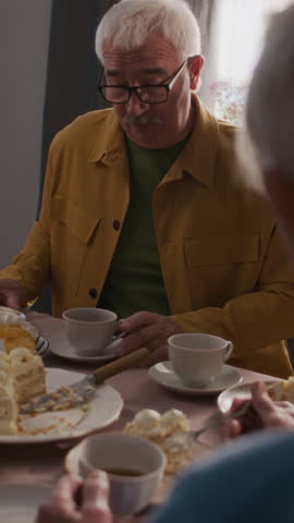 Vertical over the shoulder view of cheerful elderly man wearing glasses and mustard-coloured shirt having conversation with friend sitting at table during tea party