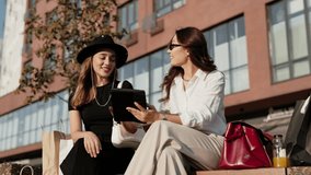 Charming women sitting outdoors near modern building. Holding shopping bags beside. Female in black hat smiling while other in sunglasses showing tablet device. Watching content together. - Powered by Shutterstock - Get 15% off with code: PIKWIZARD15