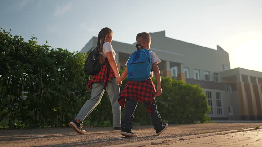 Children with backpacks walking side by side. Girl leads younger boy on road to school. Early morning walk with sibling students. School journey with backpack child walk in morning road setting