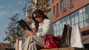 Elegant Caucasian woman wearing sunglasses and white shirt. Sitting outdoors near building. Holding tablet in hands and reading attentively. Shopping bags and red handbag placed beside while relaxing. - Powered by Shutterstock - Get 15% off with code: PIKWIZARD15