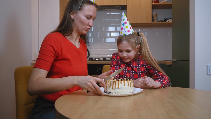 Mother in red t-shirt and party hat joyfully cutting birthday cake alongside daughter wearing plaid shirt, sharing loving moment in family kitchen during festive celebration