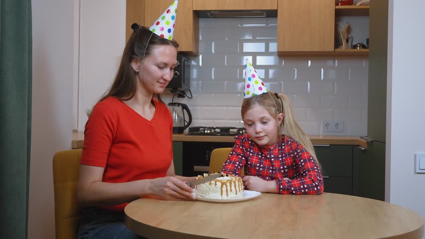 Mother in red t-shirt and party hat joyfully cutting birthday cake alongside daughter wearing plaid shirt, sharing loving moment in family kitchen during festive celebration