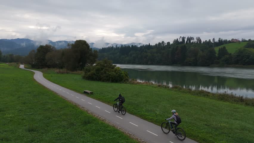 Cyclists ride on e-bikes along the Drava River through green countryside as the drone orbits left, aerial.