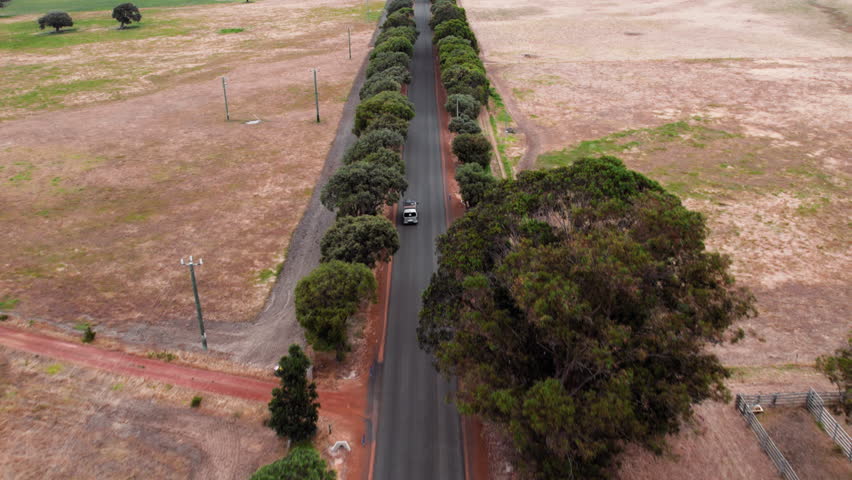 Aerial view of a camper van car drives along a tree-lined road in a wide, open area under a clear sky during the day in Australian outback