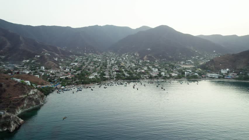 A cinematic aerial panoramic view panning across the picturesque Taganga bay, showcasing the vibrant fishing village, the many colorful local boats, and the arid foothills of the Sierra Nevada.