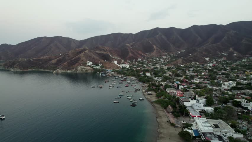 Cinematic boom shot lifting over the picturesque Taganga Bay, Colombia. Features a dense cluster of fishing boats and the coastal village nestled against the rugged Sierra Nevada mountains.