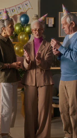 Vertical view of elegant elderly woman using smartphone with two friends standing nearby when her husband bringing cream cake with lit candles during birthday party