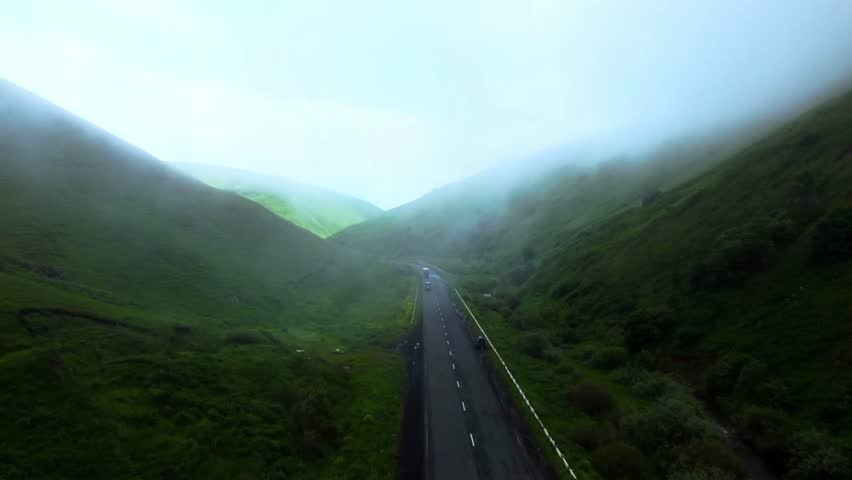 A winding road cuts through lush green hills covered in mist, creating a serene and atmospheric mountain landscape.