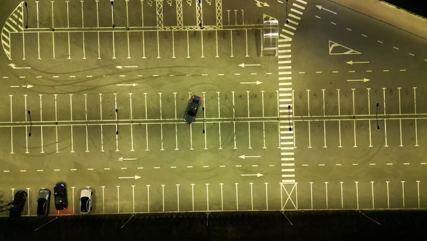 Drone descending over illuminated parking lot with drifting car at night