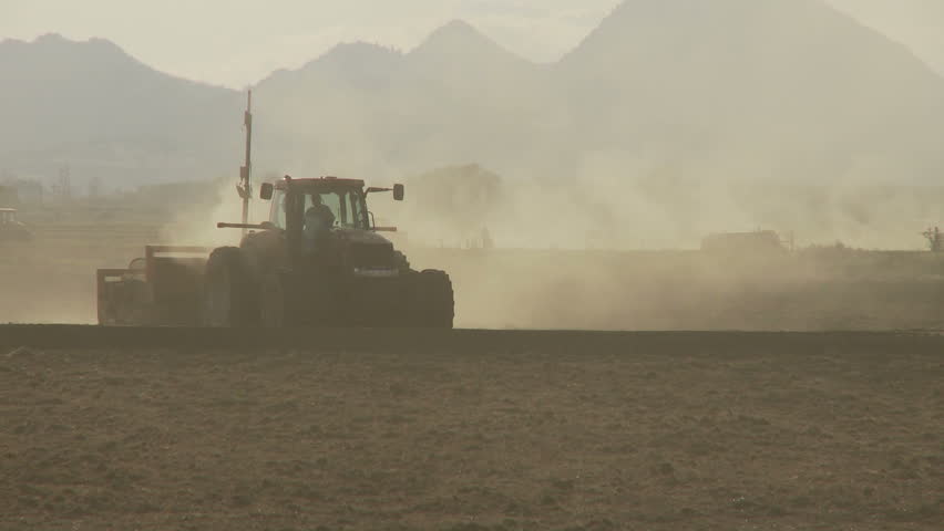 Silhouetted tractor drives through dusty field with man walking in the