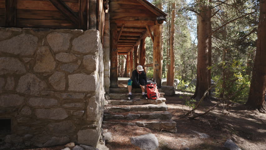 A dedicated hiker carefully adjusts their equipment while sitting on the steps of a charming cabin, which is surrounded by a tranquil and serene forest, inviting exploration
