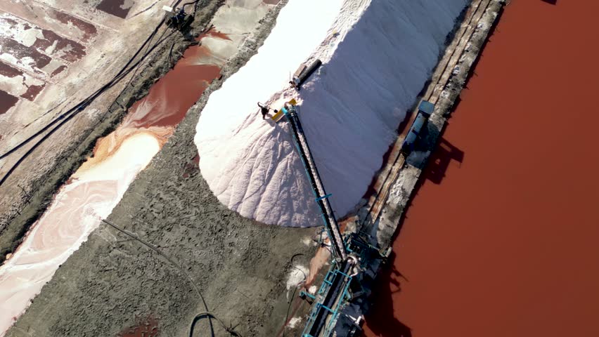 Aerial drone view of a worker harvesting pink salt with shovel near conveyor belt on large salt pile beside red evaporation pond, showing industrial mineral extraction and traditional salt production
