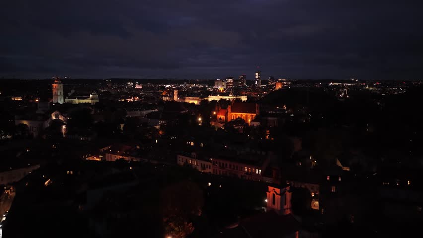 Aerial night view of Vilnius, Lithuania, featuring the illuminated Cathedral Square, Bell Tower, and modern city skyline in the background.