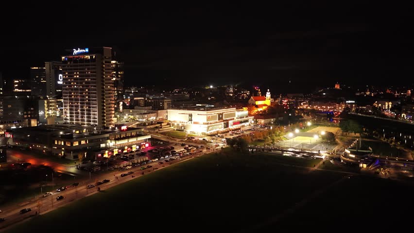 Aerial night view of Vilnius business district, featuring illuminated skyscrapers, modern glass buildings, and vibrant city lights along Konstitucijos Avenue.