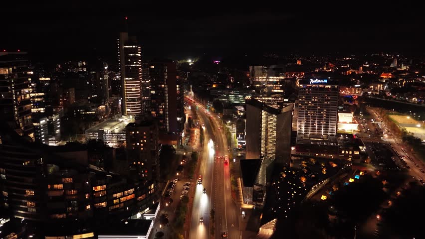 Aerial night view of Vilnius business district, featuring illuminated skyscrapers, modern glass buildings, and vibrant city lights along Konstitucijos Avenue.