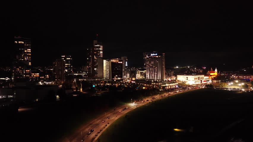 Aerial night view of Vilnius business district, featuring illuminated skyscrapers, modern glass buildings, and vibrant city lights along Konstitucijos Avenue.