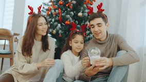 Family enjoys holiday evening by the Christmas tree while looking at a snow globe in a cozy living room decorated with ornaments - Powered by Shutterstock - Get 15% off with code: PIKWIZARD15
