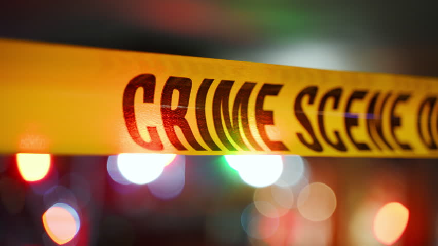 Close up of bright yellow Crime Scene tape across an urban environment, marking a restricted area for investigation. Police flashing lights are on the background