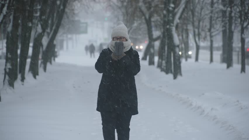 Woman tries to keep warm during snowfall in city. Female person rubs herself with mittened hands and walks through snow during winter snowstorm, wrapped in warm scarf.