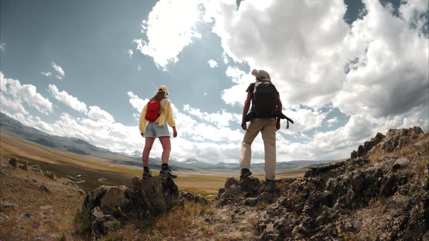 A couple of tourists male and female stand on the top of a mountain and triumphantly raise their hands up, rejoicing of achievement their goal and admiring beautiful mountain scenery. Low angle view