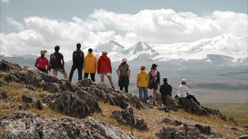 Big group of young diverse hikers are standing against snow capped mountain at winner poses with open arms and enjoys great view