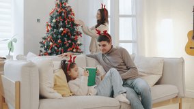 Father and daughter sit on a cozy sofa reading a book together, while the mother decorates the Christmas tree in a bright, festive living room filled with holiday cheer - Powered by Shutterstock - Get 15% off with code: PIKWIZARD15
