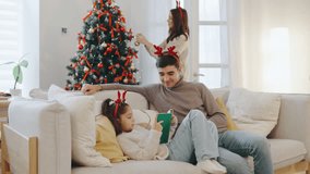 Dad and daughter wear festive antlers while reading a book on the sofa, as mom decorates the Christmas tree nearby in a cozy, cheerful holiday home - Powered by Shutterstock - Get 15% off with code: PIKWIZARD15
