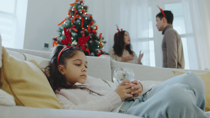 A sad girl in festive antlers sits on the sofa, gazing at a snow globe, while her parents argue near the Christmas tree in a bright, decorated room