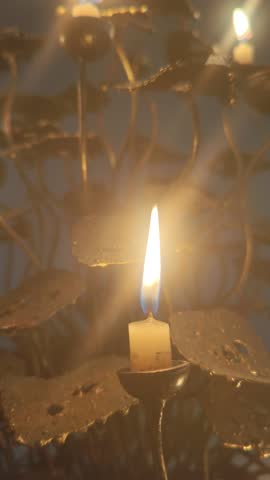 Candle flame with decorative metallic holder
A single lit candle casting a warm, steady flame that illuminates a decorative metallic holder with leaf‑like motifs and curving stems, producing soft high